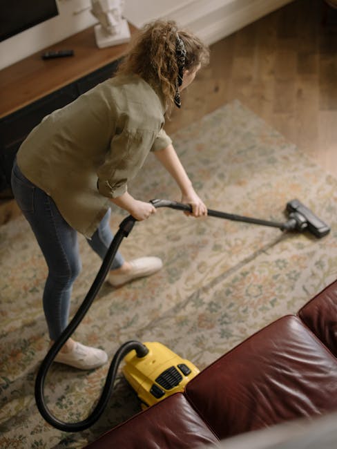 A woman with curly hair, wearing a beige jacket, blue jeans, and white shoes, is using a yellow vacuum cleaner to deep clean a patterned area rug on a wooden floor in a living room. The room features a dark brown leather sofa and a wooden sideboard with a white decorative item, with soft lighting illuminating the space. Carpet Cleaning Mayfair logo and website details suggest professional surface cleaning services specializing in domestic and commercial hygiene maintenance.