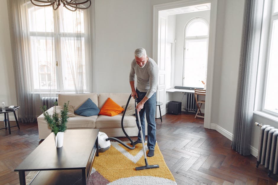 A middle-aged man with gray hair and glasses is engaged in deep cleaning a living room on Mount Street, Mayfair. He is wearing a light gray long-sleeve shirt and dark trousers, operating a vacuum cleaner on a patterned yellow, gray, and white area rug. The room features a cream-colored sofa adorned with blue, orange, and beige cushions, positioned against large windows with sheer curtains that allow natural light to fill the space. There is a dark wooden coffee table with a small potted green plant, a side table with a glass of water, and a desk with a chair near the window. The flooring consists of polished hardwood arranged in a herringbone pattern, and the room’s walls are painted white, creating a bright and airy atmosphere. The scene exemplifies professional domestic cleaning, emphasizing surface cleaning and sanitisation, and highlights the expertise of Carpet Cleaning Mayfair in maintaining a hygienic living environment.