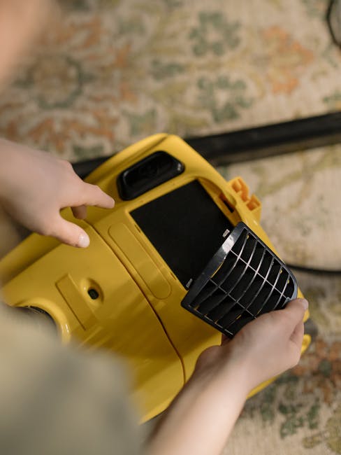 A woman with curly hair, wearing a beige jacket, blue jeans, and white shoes, is using a yellow vacuum cleaner to deep clean a patterned area rug on a wooden floor in a living room. The room features a dark brown leather sofa and a wooden sideboard with a white decorative item, with soft lighting illuminating the space. Carpet Cleaning Mayfair logo and website details suggest professional surface cleaning services specializing in domestic and commercial hygiene maintenance.