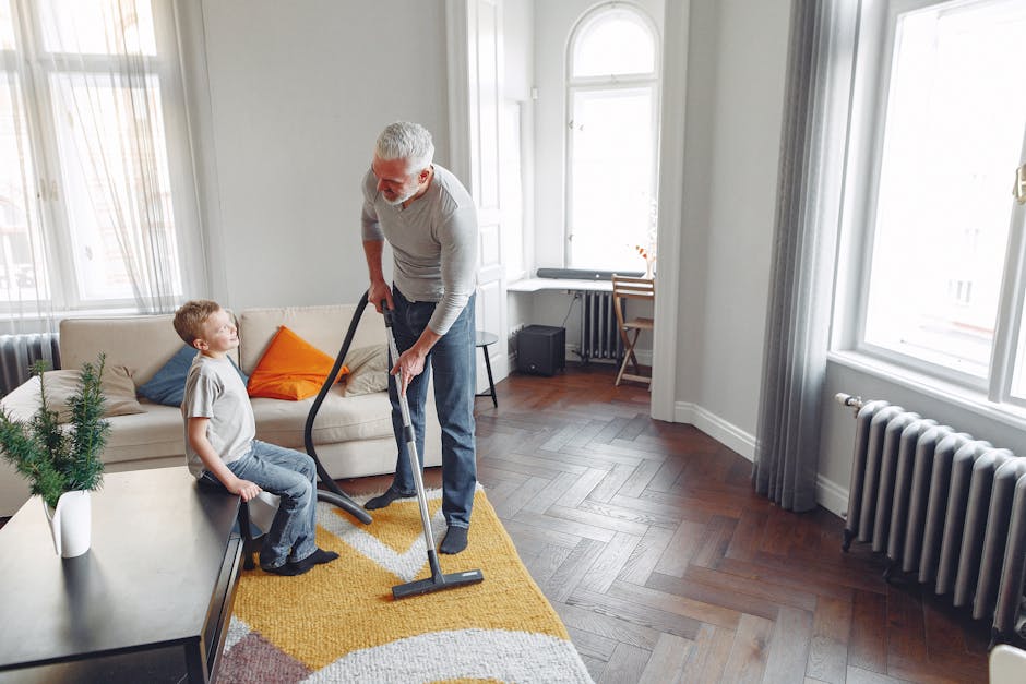 A senior man with white hair and a beard uses a vacuum cleaner to clean a yellow and white patterned area rug in a bright living room. A young boy with short hair, wearing a grey T-shirt and jeans, is seated on a beige sofa with colorful cushions in shades of orange and blue, watching the man. The room features wooden flooring, large windows with sheer and grey curtains allowing natural light, a radiator beneath one window, and a small wooden desk with a chair near the window. Visible cleaning tools include the vacuum cleaner, and the room appears tidy and well-maintained, highlighting household surface cleaning and domestic hygiene as part of Carpet Cleaning Mayfair’s services.