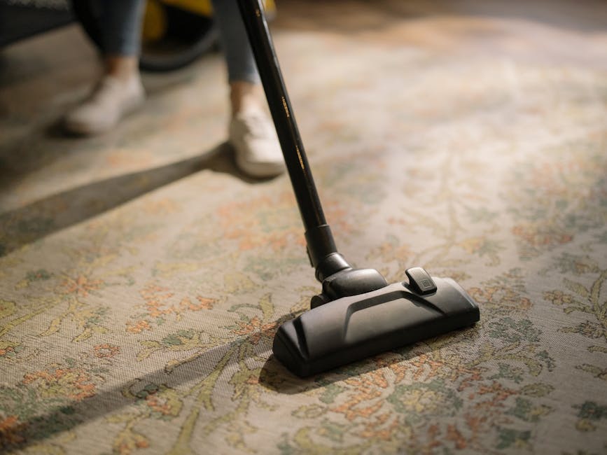 A close-up of a vacuum cleaner head actively cleaning a patterned area rug in a well-lit living room. The rug features intricate floral motifs in muted shades of beige, green, and orange, with visible dust and dirt accumulating in the fibers. The vacuum cleaner's black head and adjustable wand are partially visible, with the operator’s white shoes and dark pants seen in the background standing on the rug. The scene highlights surface cleaning and deep cleaning processes performed by Carpet Cleaning Mayfair, ensuring hygienic and well-maintained residential spaces in Berkeley Square.