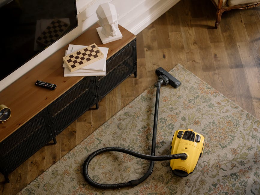 A yellow canister vacuum cleaner with a flexible hose and cleaning head resting on a patterned area rug in a living room. The room features wooden flooring, a black metal TV stand with a flat-screen TV, a remote control, and a stack of papers with a chessboard book on top. On the TV stand, there's a white decorative bust, and part of a brown upholstered armchair is visible in the corner. The scene is well-lit, highlighting the clean surfaces and the tidy appearance of the space, with attention to surface cleaning and maintenance in a domestic setting. Carpet Cleaning Mayfair provides professional deep cleaning and surface sanitisation, maintaining hygiene standards within residential areas.