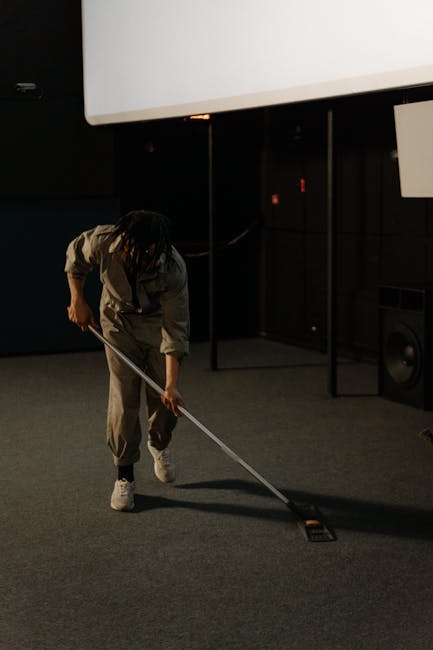 A person operating a yellow portable carpet cleaning machine with a black grille on top, positioned on a traditional patterned rug with muted tones. The individual’s hands are visible, holding the d
