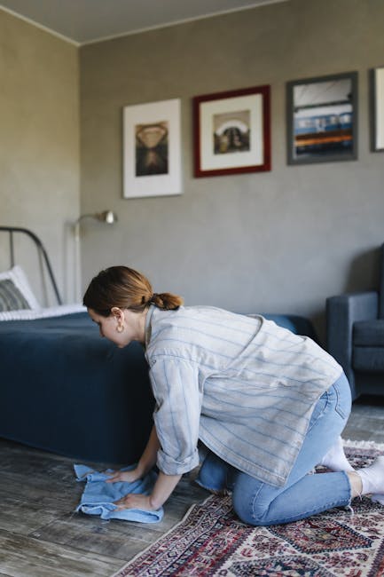 A woman with brown hair tied back, wearing a light blue and white striped shirt and jeans, kneeling on a patterned rug and cleaning a dark wooden floor with a blue cloth. The room features a gray wall