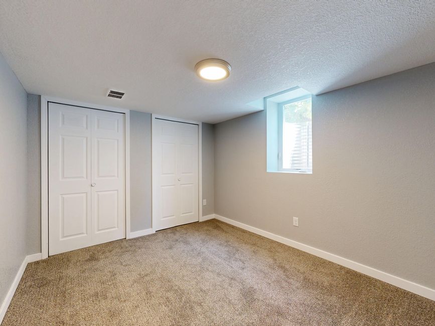 A small, clean basement room with light grey painted walls and a beige, textured carpeted floor. The room features two white, closed double doors for storage or closet space on the left side, with simple round knobs. There is a small rectangular window on the right wall, allowing natural light to enter the space, and a modern, flush-mounted ceiling light in the center providing additional illumination. The room appears well-maintained, free of dust or clutter, with neat white baseboards along the bottom of the walls. Visible air vent near the ceiling adds to the ventilation. Carpet Cleaning Mayfair's services focus on surface cleaning, deep cleaning, and sanitisation to maintain hygienic conditions in residential spaces like this basement.