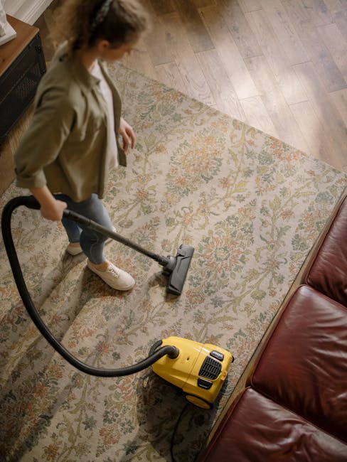 A person using a yellow vacuum cleaner with a black hose and a cleaning head attachment to perform surface cleaning on an ornate, patterned area rug with floral motifs in beige, green, and orange hues
