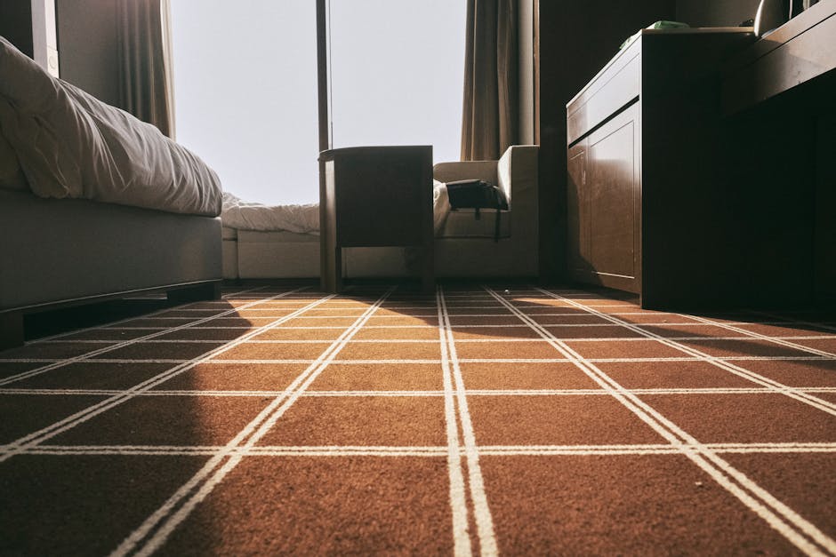 A woman with curly brown hair is kneeling on a wooden floor, carefully lifting the edge of a woven jute rug to reveal the polished hardwood surface underneath. The room features a light grey upholster