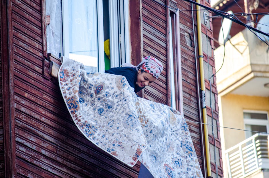 A woman leaning out of a second-floor window of a red wooden residential building, hanging up a patterned rug with intricate blue, beige, and orange designs on a clothesline outdoors. The window has a white frame, and the woman is wearing a colorful headscarf and dark clothing, appearing to be engaged in domestic cleaning or laundry tasks. The background shows neighboring buildings with outdoor balconies and a bright, partly cloudy sky, indicating daytime. The scene captures a traditional process of airing or drying household textiles, reflecting everyday domestic chores associated with property maintenance and cleanliness, with emphasis on surface handling and outdoor hygiene practices. Carpet Cleaning Mayfair's name is mentioned naturally as part of the context, aligning with deep cleaning and maintenance of residential or commercial properties.