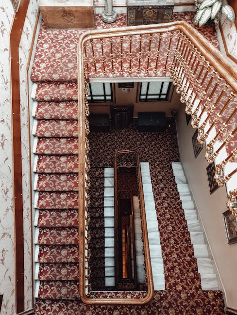Aerial view of an ornate multi-story staircase in a classic Victorian interior, featuring a patterned red and gold carpet on each flight, with polished wooden banisters and golden balustrades. The staircase connects three levels, with the top landing visible, decorated with framed artwork on the walls and a large potted plant in the corner. Natural light illuminates the space, highlighting the intricate details of the woodwork and the plush carpet, indicating a well-maintained, clean environment. The overall setting reflects a historic residential or commercial building, with a focus on surface cleaning and preservation. Carpet Cleaning Mayfair's expertise in deep cleaning and maintaining such elegant surfaces is essential for keeping this grand staircase in top condition.